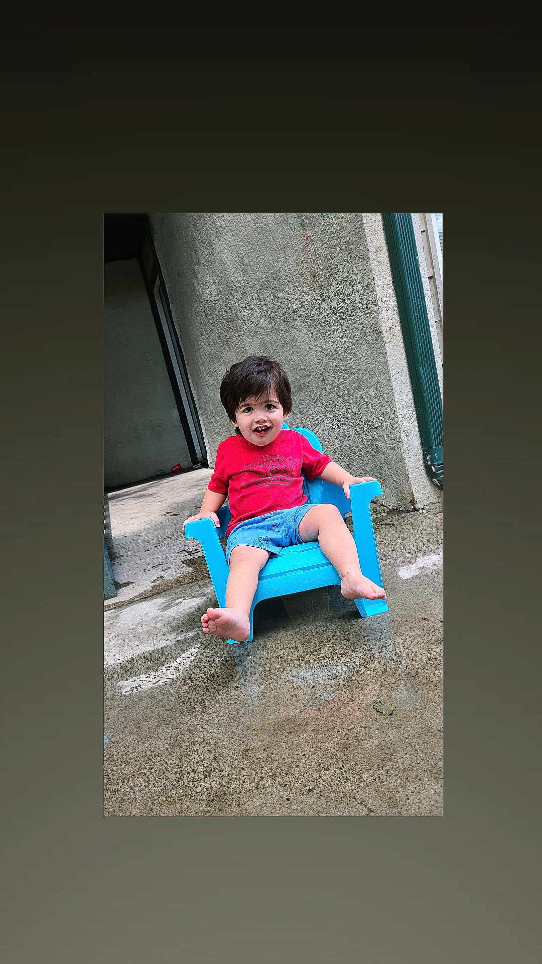 Zayne is registered to the contest to win money with this photo: barefoot, blue_chair, blue_shorts, casual_clothing, child, concrete_floor, daylight, happy, outdoor, person, playful, portrait, red_shirt, relaxed, short_hair, sitting, smiling, toddler, wall, young_child
