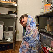 Zayne is registered to the contest to win money with this photo: child, blanket, kitchen, pantry, wooden_floor, smiling, happy, indoors, shelf, container, food_storage, metal_shelf, person, floor, cozy, young, cute, wrapped, face, casual