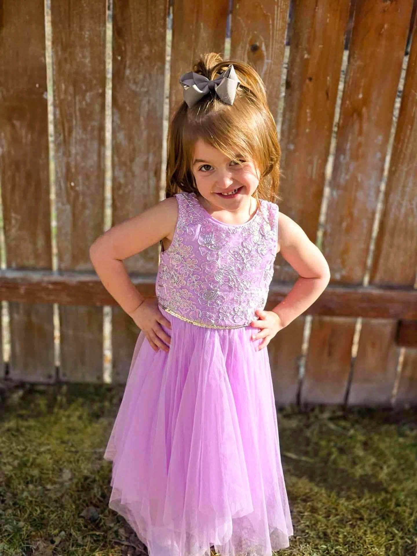 Jazlyn is registered to the contest to win money with this photo: child, girl, dress, purple_dress, bow, smiling, outdoor, sunlight, grass, fence, happy, pose, flower_embroidery, young, person, portrait, fashion, cute, spring, playful