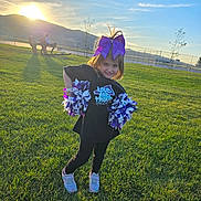 Jazlyn is registered to the contest to win money with this photo: child, girl, cheerleader, pom_poms, purple_bow, smiling, outdoor, grass, sunset, field, casual_clothing, playful, sunlight, nature, young_child, shoes, posing, happy, recreation, daytime