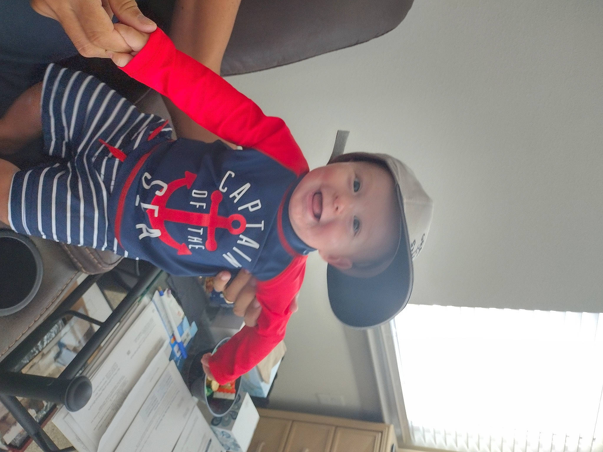 Tyson is registered to the contest to win money with this photo: baby, baby_toddler_clothing, baseball_cap, cap, child, elbow, fun, gesture, hat, headgear, headwear, human_leg, knee, person, room, sitting, smile, sneakers, t_shirt, toddler