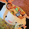 baby, bunny_ears, celebration, child, colorful, cushion, cute, decorations, easter_basket, easter_eggs, festive, holiday, indoors, infant, lying_down, pink_socks, plush, smiling, soft_texture, toy