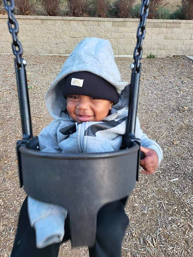 King is registered to the contest to win money with this photo: baby, baby_carriage, baby_products, baby_toddler_clothing, child, city, facial_expression, fun, grass, hand, headwear, human_settlement, leisure, person, plant, playground, recreation, sitting, smile, swing