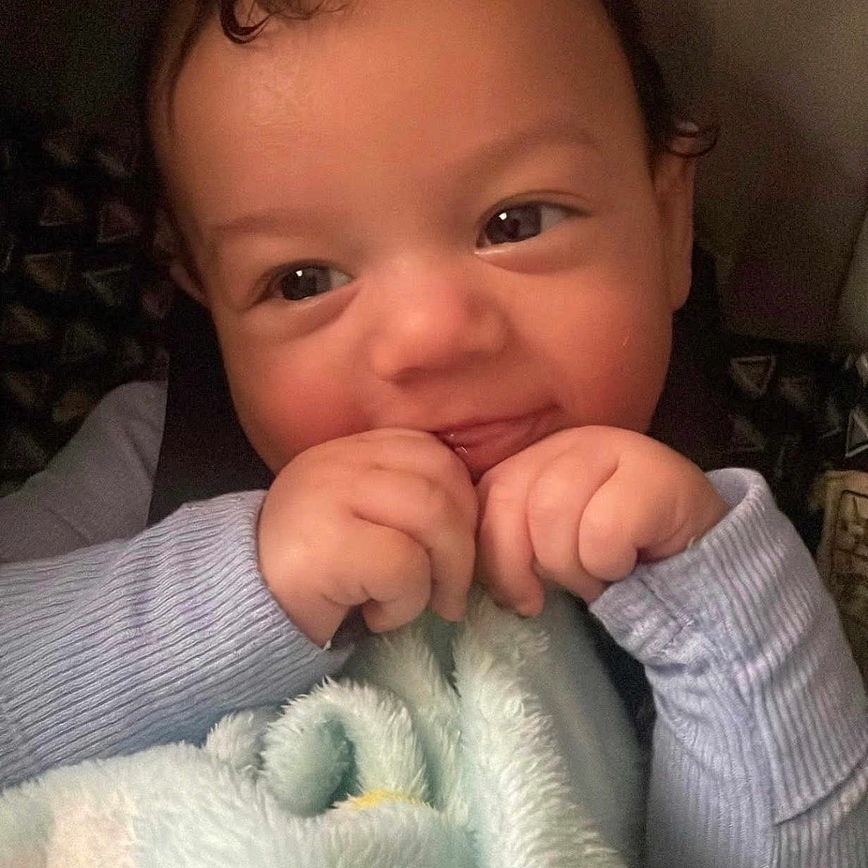 Zamir is registered to the contest to win money with this photo: baby, child, face, hands, blanket, curly_hair, smiling, blue_clothing, infant, cute, cozy, soft_texture, indoors, portrait, closeup, expression, skin, person, toddler, warm