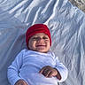 baby, smiling, red_hat, red_pants, white_shirt, lying_down, white_sheet, sand, outdoor, sunlight, child, happy, infant, clothing, headwear, portrait, relaxed, cute, one_person, casual