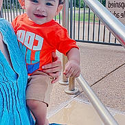 Wahaj joined the competition — help win amazing prizes! adult_hand, blue_jeans, blue_top, child, clouds, curly_hair, daytime, happy, holding, orange_shirt, outdoor, person, portrait, railing, shorts, sidewalk, sky, smiling, sunny, toddler