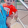 adult_hand, blue_jeans, blue_top, child, clouds, curly_hair, daytime, happy, holding, orange_shirt, outdoor, person, portrait, railing, shorts, sidewalk, sky, smiling, sunny, toddler