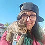 Two is registered to the contest to win money with this photo: animal, brown, cap, casual, cat, clothing, daylight, glasses, green_hair, hair, leash, outdoor, person, pet, portrait, shoulder, sky, smile, tabby_cat, urban