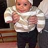 baby, child, smiling, striped_sweater, hat, sneakers, indoor, adult_hands, table, red_tablecloth, black_pants, cheerful, portrait, person, home, furniture, photography, cute, happy, standing