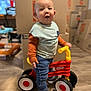 cardboard_boxes, casual_clothing, child, curious, expression, face, flooring, furniture, happy, home, indoor, jeans, long_sleeve_shirt, moving, person, playtime, sneakers, standing, toddler, toy_bike