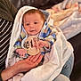 baby, infant, newborn, one_month, milestone_sticker, blanket, couch, adult_hand, baby_hand, hoodie, plaid_jacket, striped_shirt, jeans, portrait, indoor, cozy, bokeh_background, eyes, cheeks, skin