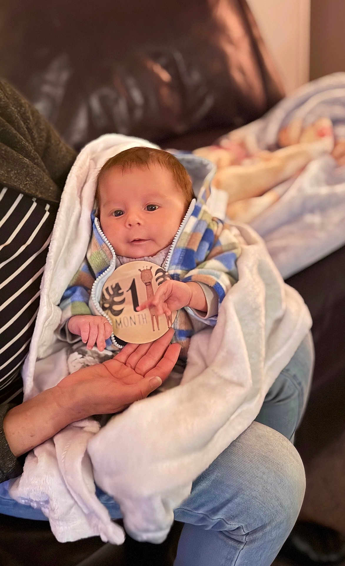 Gabriel joined the competition — help win amazing prizes! baby, infant, newborn, one_month, milestone_sticker, blanket, couch, adult_hand, baby_hand, hoodie, plaid_jacket, striped_shirt, jeans, portrait, indoor, cozy, bokeh_background, eyes, cheeks, skin