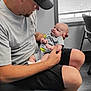 Dominic is registered to the contest to win money with this photo: man, baby, indoor, window, blinds, cap, sunglasses, onesie, seated, holding, floor, chair, tattoo, shorts, sandals, adult, child, affection, interaction, casual_clothing