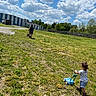 Elianna is registered to the contest to win money with this photo: child, toddler, adult, grass, field, sky, clouds, toy_lawn_mower, lawn_mower, greenery, outdoor, nature, summer, casual_clothing, shorts, tshirt, boots, trees, fence, building