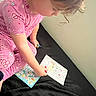 child, pajamas, pink_clothing, coloring, cartoon, paper, stickers, curly_hair, bed, black_bedspread, indoor, activity, person, young_child, art, drawing, concentration, cute, play, fun
