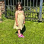 Laila is registered to the contest to win money with this photo: child, girl, yellow_dress, pink_sneakers, grass, garden, iron_gate, brick_wall, sunlight, smiling, standing, outdoor, greenery, summer, portrait, person, happy, casual, nature, fashion