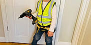 Feroze is registered to the contest to win money with this photo: child, smiling, indoor, wooden_floor, white_door, yellow_safety_vest, toy_power_drill, blue_jeans, brown_boots, construction_theme, person, standing, happy, play, pretend, young, home, casual_clothing, flooring, door_handle