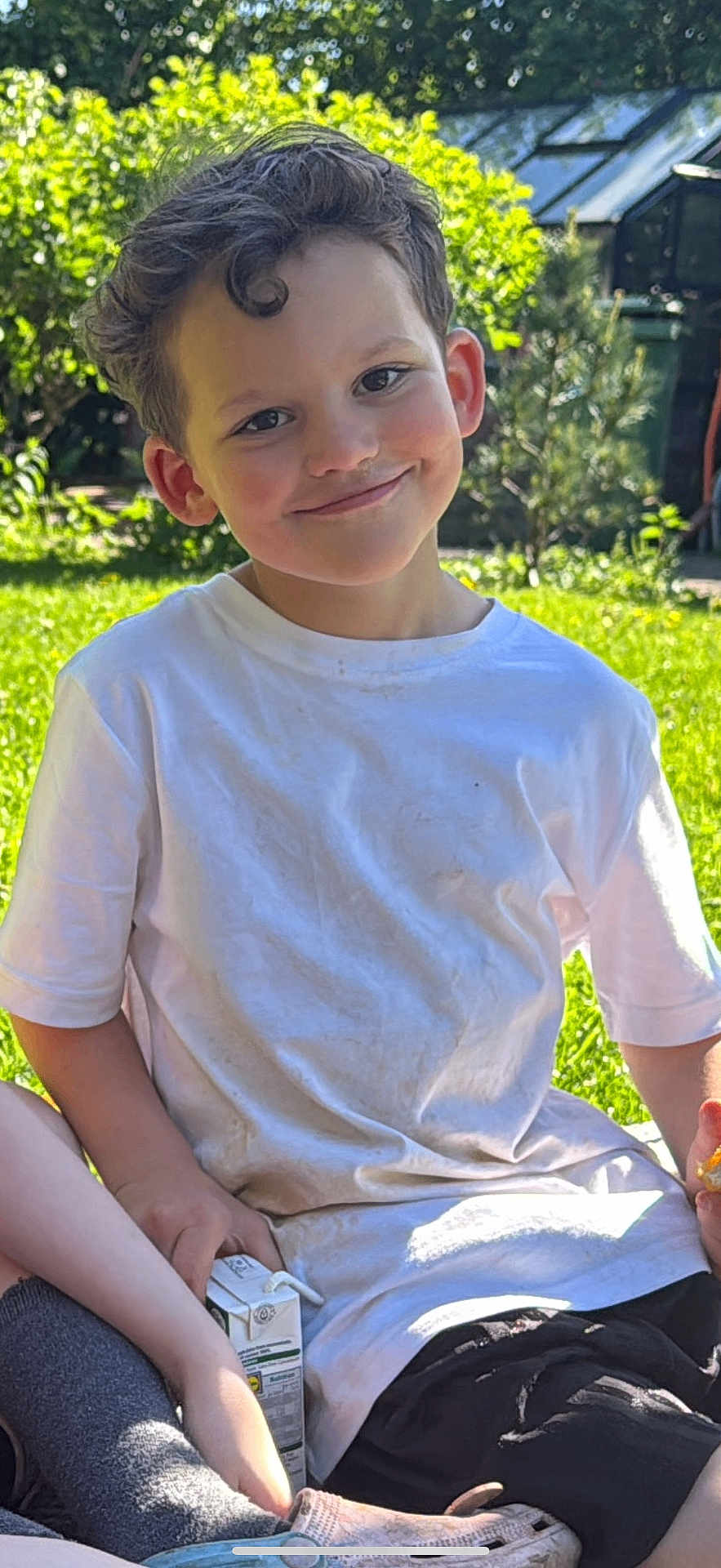 Feroze joined the competition — help win amazing prizes! child, boy, smiling, outdoor, grass, white_tshirt, curly_hair, juice_box, snack, sunlight, garden, greenery, casual_clothing, happy, sitting, portrait, daylight, nature, relaxed, playful