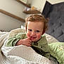 toddler, child, smile, blanket, bedroom, cozy, green_clothing, face, rosy_cheeks, bed, pillow, curly_hair, indoor, soft_light, cute, happy, person, portrait, furniture, relaxing