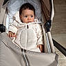 baby, infant, stroller, beige, coat, padded, indoor, floor, curious, child, portrait, warm_clothing, seat, young_child, cute, soft_light, face, hands, expression, blanket