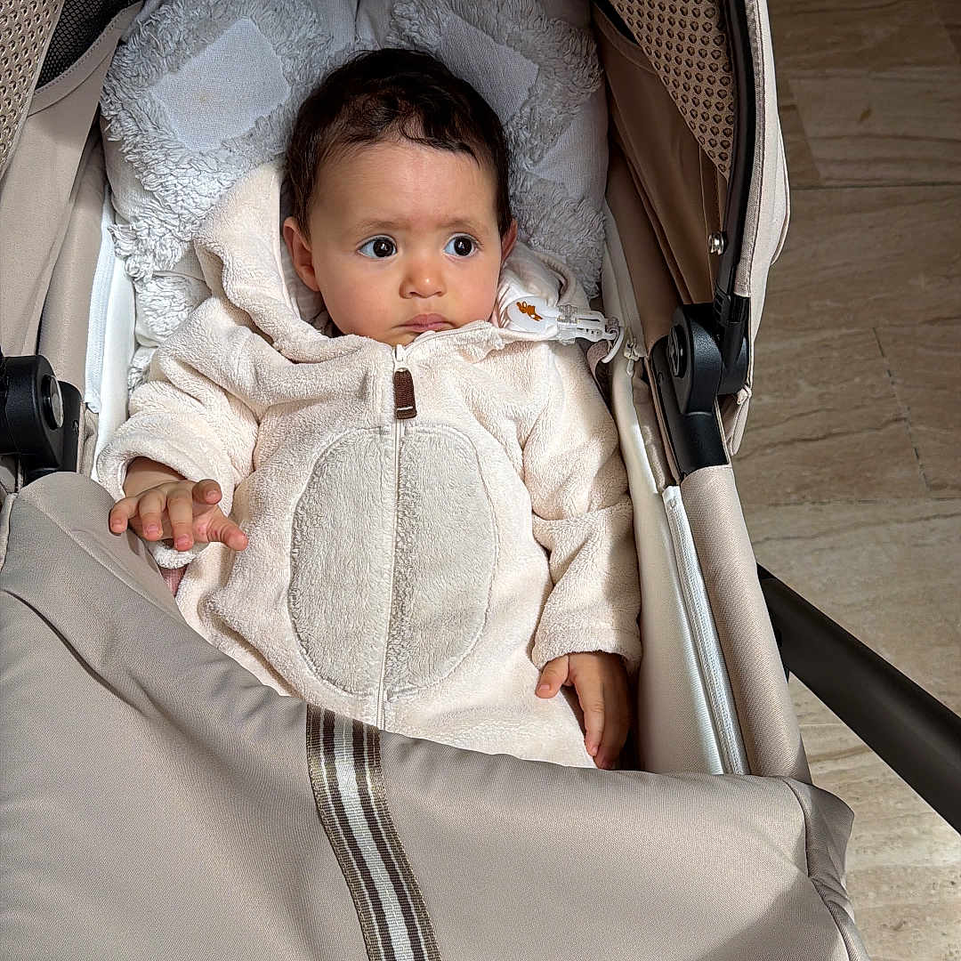Rita participe au concours pour gagner de l'argent avec cette photo : baby, infant, stroller, beige, coat, padded, indoor, floor, curious, child, portrait, warm_clothing, seat, young_child, cute, soft_light, face, hands, expression, blanket