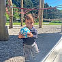 toddler, child, soccer_ball, playground, outdoor, sunny, rope, wooden_poles, climbing_structure, gravel, casual_clothing, sneakers, young_child, sports_equipment, greenery, trees, daylight, fun, activity, park
