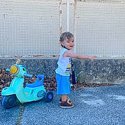 Milan participe au concours pour gagner de l'argent avec cette photo : toddler, child, toy_scooter, pointing, outdoor, pavement, fence, dry_leaves, blue_shorts, white_shirt, brown_shoes, curly_hair, playtime, side_view, daylight, smiling, casual_clothing, standing, keys, scarf