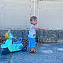 toddler, child, toy_scooter, pointing, outdoor, pavement, fence, dry_leaves, blue_shorts, white_shirt, brown_shoes, curly_hair, playtime, side_view, daylight, smiling, casual_clothing, standing, keys, scarf