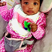 World is registered to the contest to win money with this photo: baby, infant, child, pink_clothing, bib, baby_seat, toy, curly_hair, hair_tie, seatbelt, cushion, indoor, portrait, cute, young_child, person, looking_at_camera, close_up, hand, face