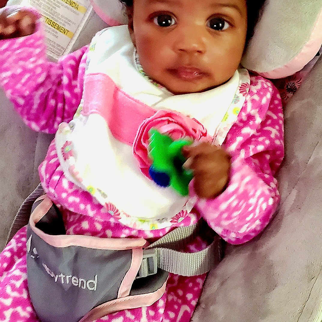 World is registered to the contest to win money with this photo: baby, baby_seat, bib, child, close_up, curly_hair, cushion, cute, face, hair_tie, hand, indoor, infant, looking_at_camera, person, pink_clothing, portrait, seatbelt, toy, young_child