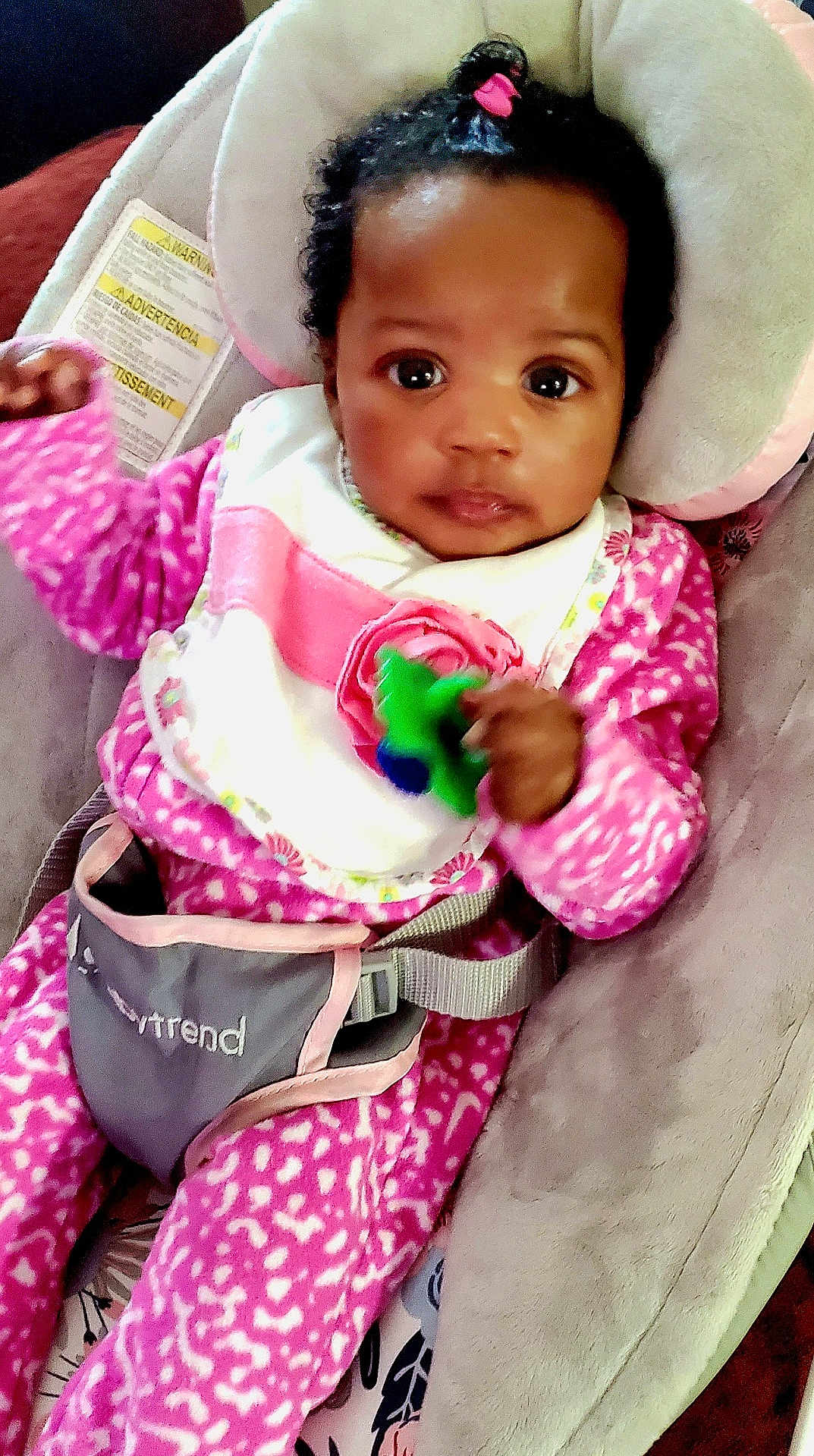 World is registered to the contest to win money with this photo: baby, infant, child, pink_clothing, bib, baby_seat, toy, curly_hair, hair_tie, seatbelt, cushion, indoor, portrait, cute, young_child, person, looking_at_camera, close_up, hand, face