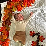 baby, infant, autumn_leaves, chestnuts, white_sheet, striped_clothing, brown_pants, socks, lying_down, curious_expression, indoor, floor_tiles, fall_season, decorative_arrangement, cute, child, small_hands, small_feet, colorful_leaves, number_three
