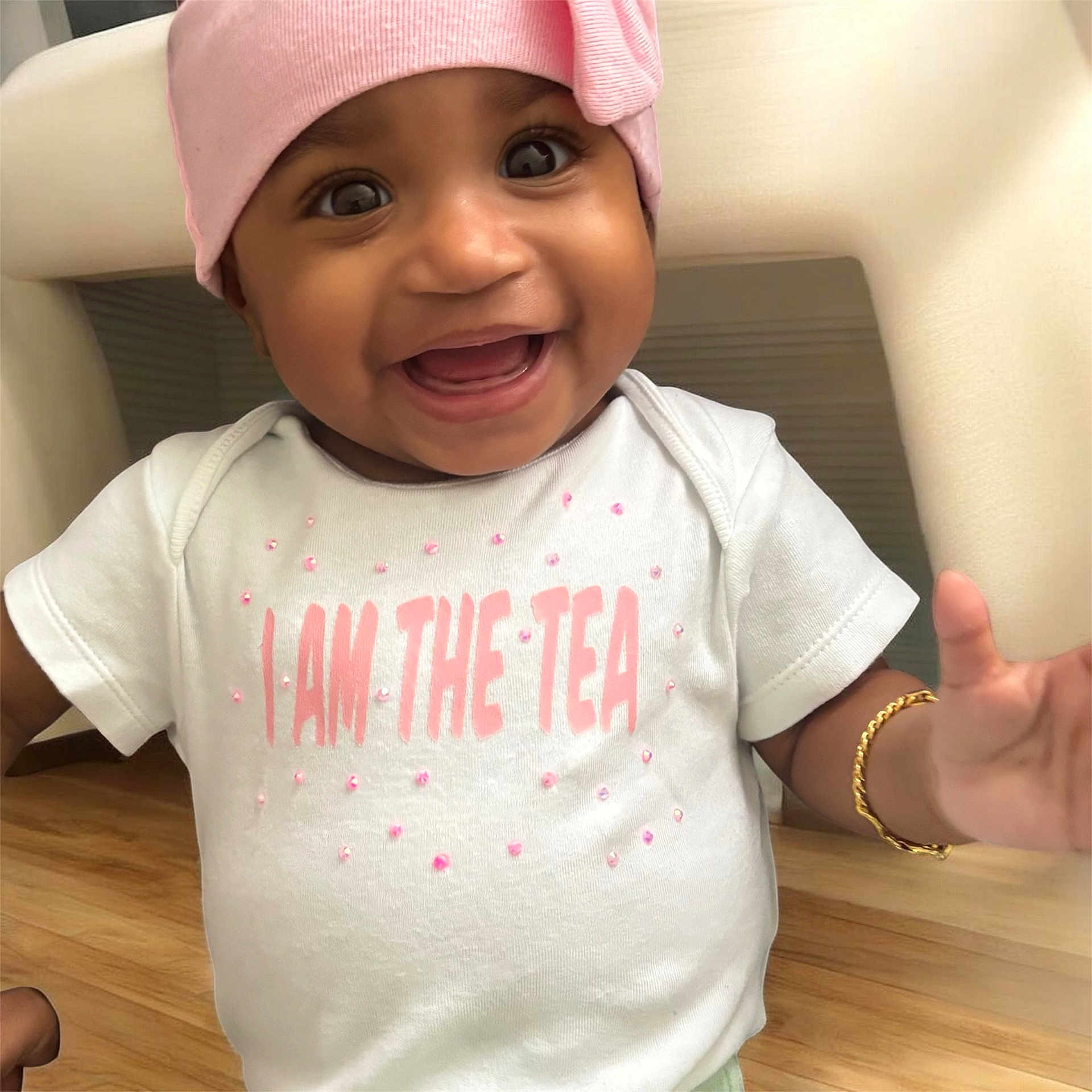 Nicole joined the competition — help win amazing prizes! baby, smiling, child, hat, pink, white_shirt, text_on_clothing, bracelet, hand, happy, indoors, floor, wooden_floor, portrait, cute, person, infant, face, apparel, sitting