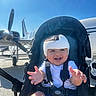 baby, child, stroller, helmet, smile, happy, outdoor, sunny, blue_sky, airplane, propeller, seatbelt, infant, transportation, daylight, person, clothing, sitting, expression, airport