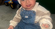 Dominic is registered to the contest to win money with this photo: baby, carpet, child, clothing, curious, denim_overalls, face, floor, furniture, hand, headgear, helmet, indoor, person, playful, room, sitting, sock, toddler, toy_vehicle