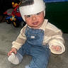 Dominic is registered to the contest to win money with this photo: toddler, child, baby, helmet, denim_overalls, sitting, carpet, toy_vehicle, indoor, playful, curious, clothing, sock, hand, face, headgear, room, furniture, floor, person