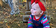 Ivy is registered to the contest to win money with this photo: baby, child, face_paint, red_headband, colorful_clothing, denim_overalls, barefoot, fall_leaves, outdoor, yard, halloween, tombstone, angel_statue, skeleton_decoration, spooky, grass, tree, house, daylight, smiling