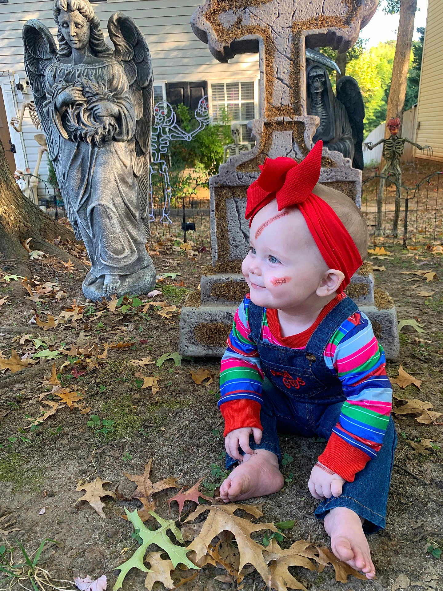 Ivy is registered to the contest to win money with this photo: baby, child, face_paint, red_headband, colorful_clothing, denim_overalls, barefoot, fall_leaves, outdoor, yard, halloween, tombstone, angel_statue, skeleton_decoration, spooky, grass, tree, house, daylight, smiling