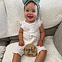 Avery joined the competition — help win amazing prizes! baby, child, smiling, headband, bow, white_outfit, couch, blanket, wooden_sign, home_interior, happy, sitting, infant, portrait, cute, indoors, person, clothing, furniture, decor