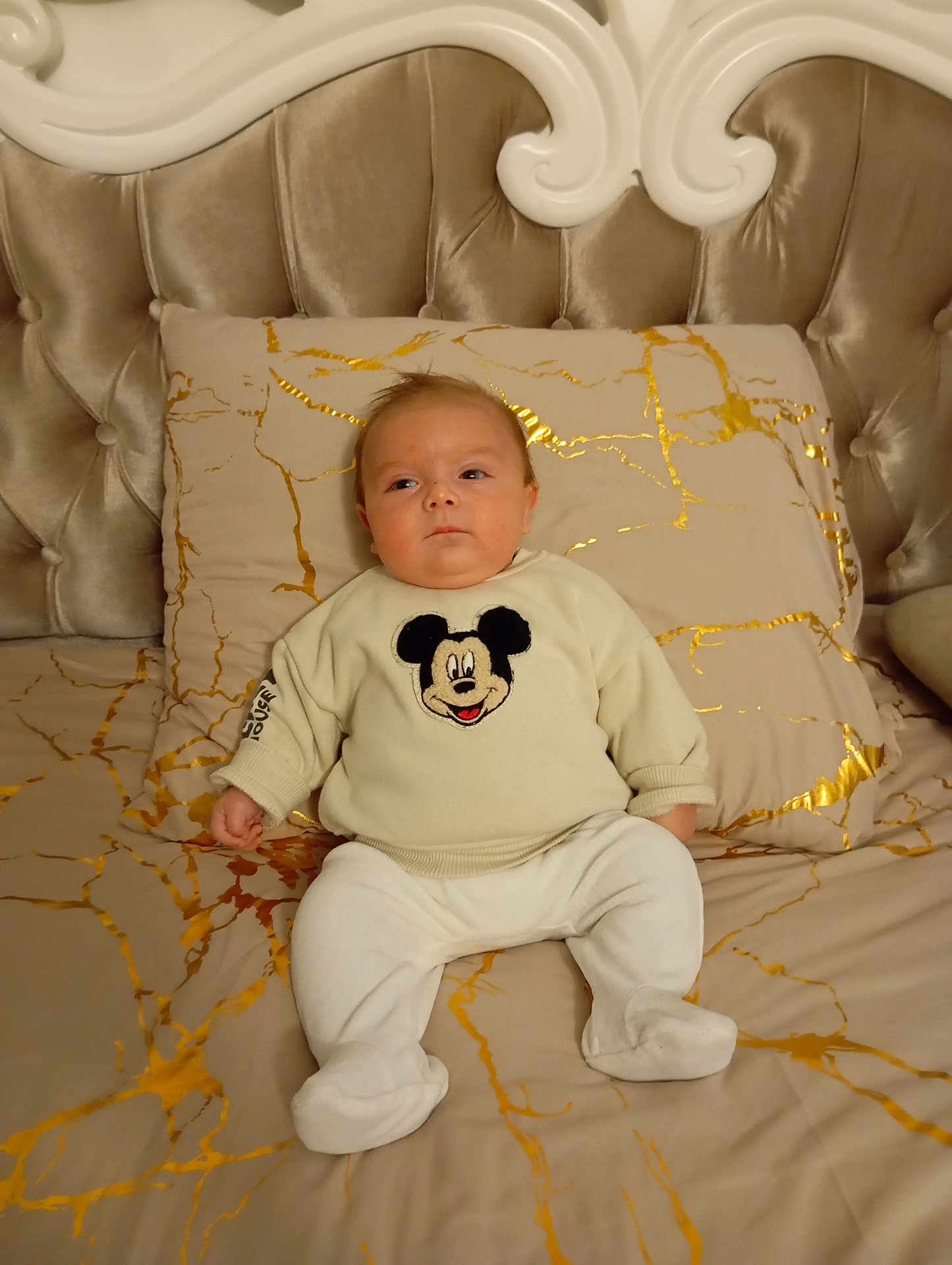 Leão participe au concours pour gagner de l'argent avec cette photo : baby, infant, bed, pillow, bedding, headboard, mickey_mouse, sweater, onesie, pajamas, portrait, sleepy, indoor, cozy, gold_pattern, tufted_headboard, blanket, sitting, face, feet