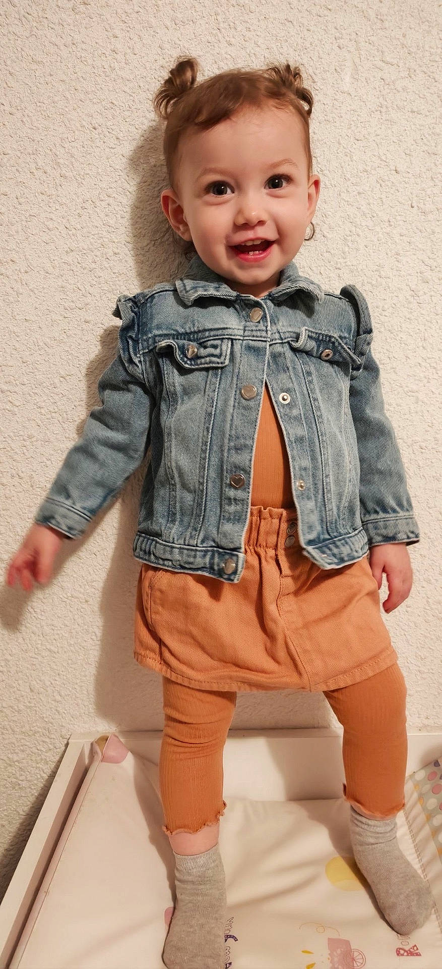 Inaya participe au concours pour gagner de l'argent avec cette photo : baby, child, cute, denim_jacket, face, fashion, hair_buns, happy, indoors, leggings, orange_clothing, person, portrait, skirt, smiling, socks, standing, toddler, wall, young_child