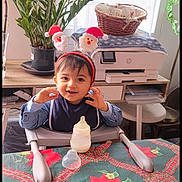 Elyes participe au concours pour gagner de l'argent avec cette photo : toddler, child, high_chair, baby_bottle, festive_headband, smile, indoor, tablecloth, poinsettia, plant, printer, basket, cupboard, table, holiday, celebration, clothing, person, face, home