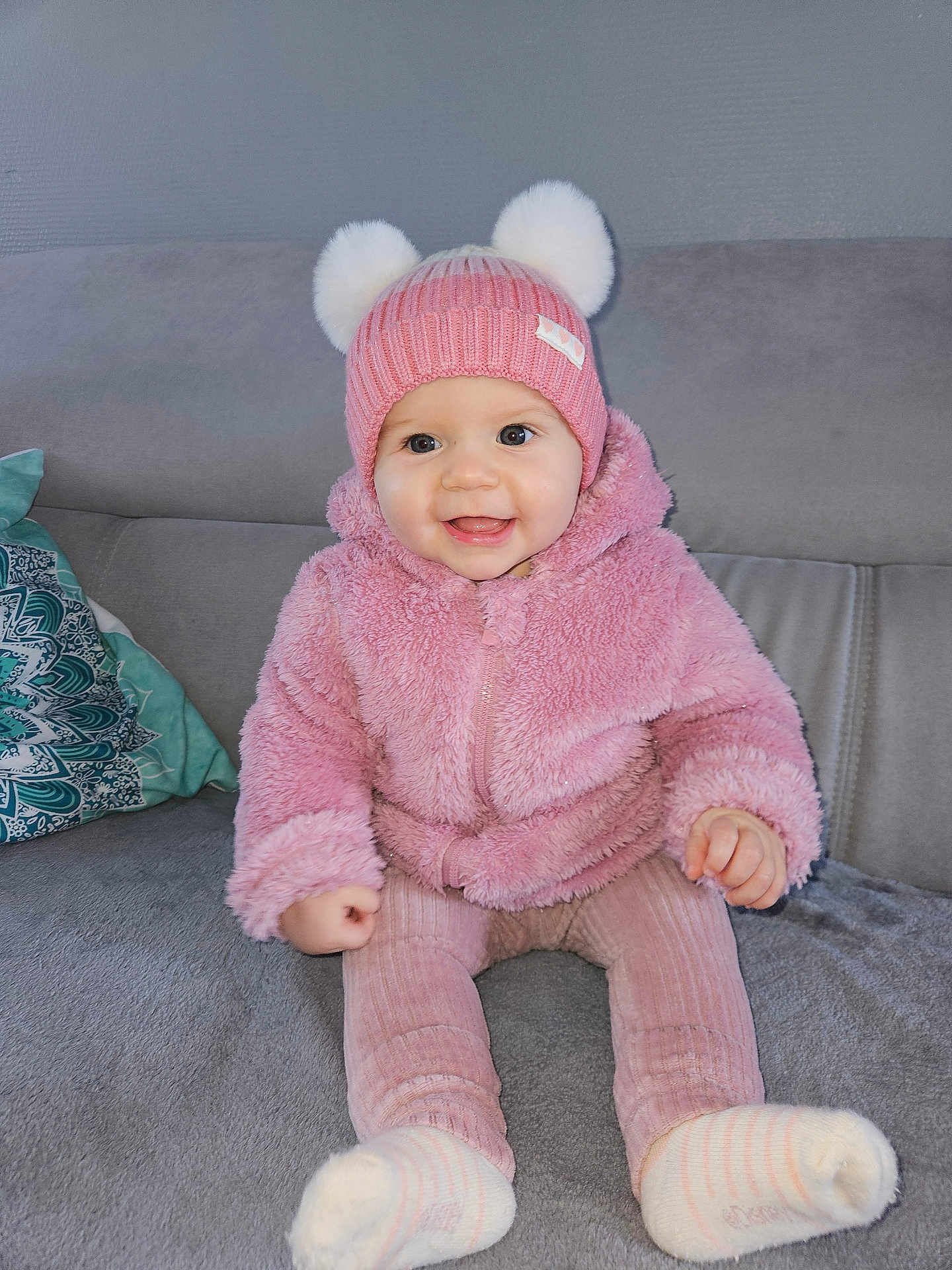 Lana participe au concours pour gagner de l'argent avec cette photo : baby, child, pink_hat, pom_poms, pink_jacket, smiling, sitting, couch, gray_sofa, cozy_clothing, socks, indoors, cute, happy, portrait, young_child, warm_clothing, person, infant, home
