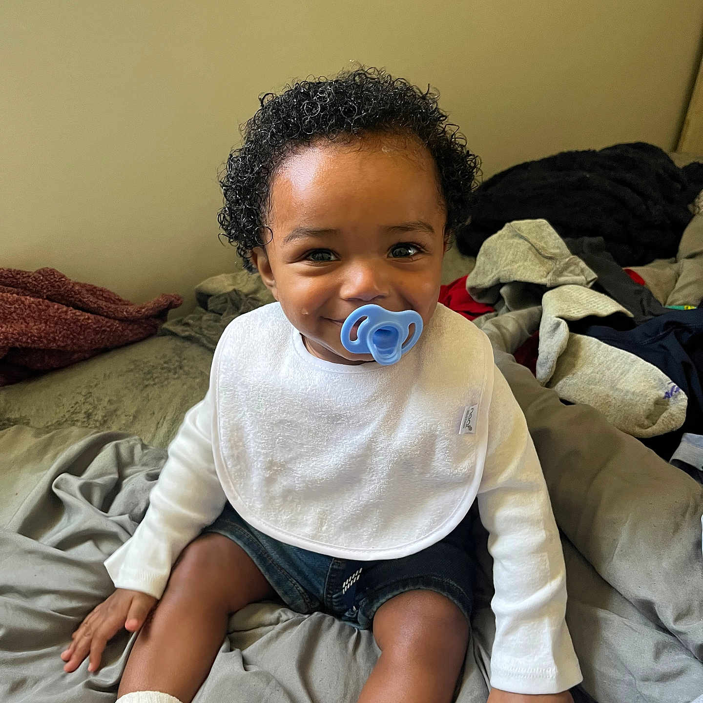 Kodell is registered to the contest to win money with this photo: baby, bed, blanket, casual_clothing, child, clothes, cozy, curly_hair, cute, denim_shorts, happy, indoor, infant, pacifier, person, sitting, smile, toddler, white_bib, white_socks