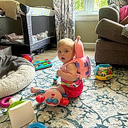 Gracelynn is registered to the contest to win money with this photo: baby, child, toy, backpack, snail, carpet, living_room, window, curtains, sofa, floor, play, indoor, person, colorful, cushion, baby_bed, plush_toy, child_sitting, daylight