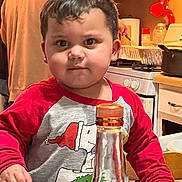 Adrian is registered to the contest to win money with this photo: background_person, bottle, cabinet, chair, child, clothing, cooking, curly_hair, expression, face, gray_shirt, hand, indoor, kitchen, person, pot, red_sleeves, stove, table, toddler