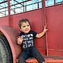 toddler, child, smile, black_clothing, cowboy_boots, red_trailer, outdoor, daylight, grass, tire, metal_structure, sitting, playful, happy, face, person, young_child, nature, casual, funny_shirt