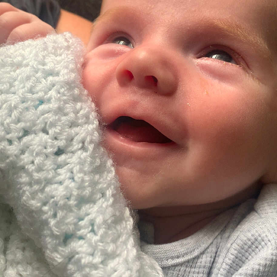 Hunter is registered to the contest to win money with this photo: baby, blanket, closeup, cute, face, happy, indoors, infant, knitted, light, mouth, natural_light, person, portrait, smile, soft_texture, toddler, white_clothing, wrapped, young_child