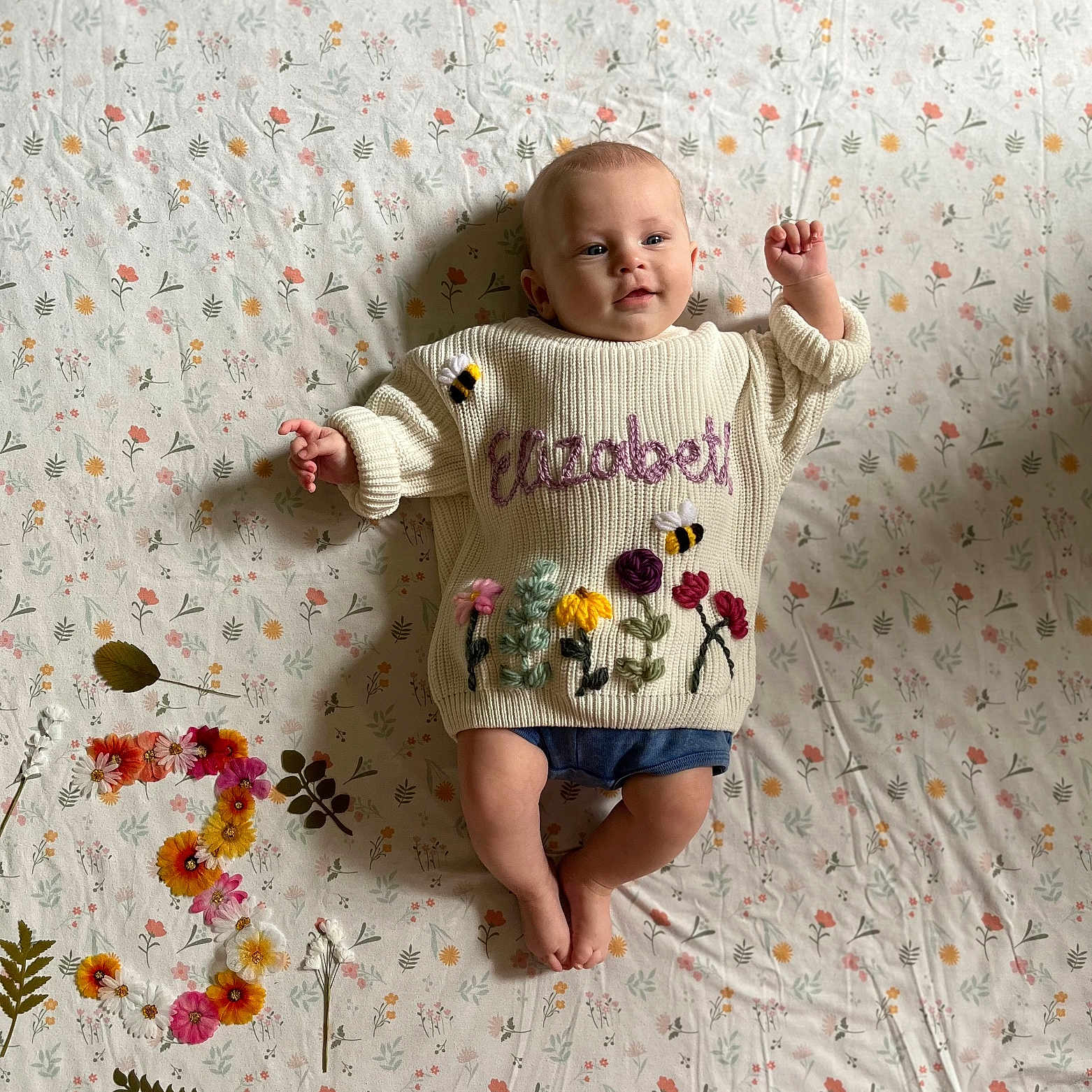 Elizabeth is registered to the contest to win money with this photo: baby, bodypart, clothing, face, finger, flower, hand, head, knitwear, newborn, pants, pattern, person, photography, plant, portrait, sitting, sleeve, sweater, t_shirt