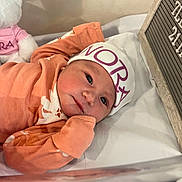 Nora is registered to the contest to win money with this photo: newborn, baby, crib, hat, orange_clothing, teddy_bear, soft_toy, face, infant, sleeping_baby, blanket, wooden_board, letter_board, lying_down, cute, portrait, indoors, resting, child, peaceful